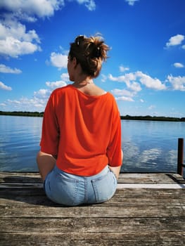 Sitting peacefully on a wooden dock, a woman gazes at the calm lake water, surrounded by a clear blue sky dotted with clouds on a beautiful day.