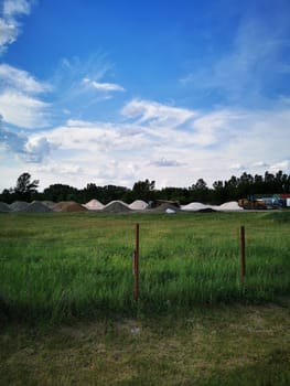 A vast construction area showcases mounds of sand and gravel, framed by lush green grass and a bright blue sky, indicating ongoing development work in the region.