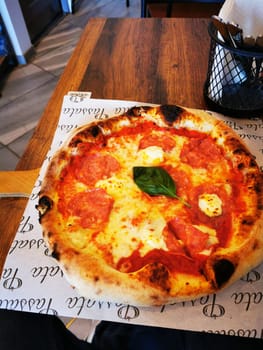 A freshly baked pizza features generous slices of pepperoni and mozzarella cheese, garnished with a sprig of basil, placed on a rustic wooden table in a casual dining environment.