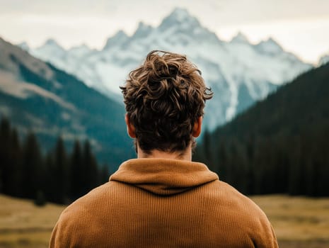 A man with curly hair is standing in a field with mountains in the background. He is wearing a brown sweater and looking out at the mountains. Concept of peace and tranquility