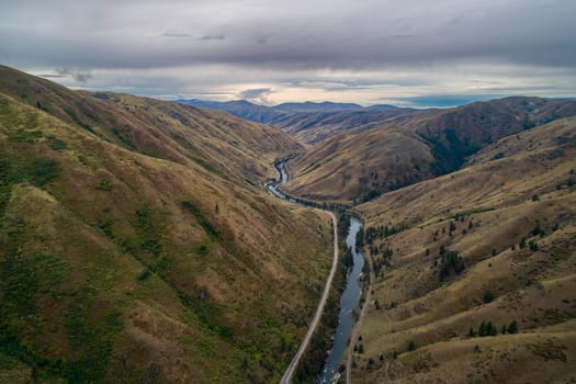 A long, winding road that runs through a valley. The road is surrounded by hills and trees, and there is a river running alongside it. The scene has a peaceful and serene mood