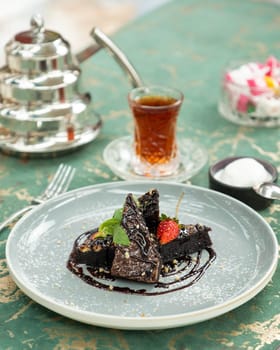 Slices of chocolate brownie are being served on a plate with chocolate sauce, a strawberry, nuts, powdered sugar and mint with a glass of tea and turkish delight in the background