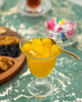 Glass of quince dessert is sitting on a table with a spoon in it, next to a plate of dried fruits and a cup of turkish tea