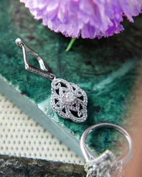 Silver earrings and ring with diamonds are lying on a green malachite stone next to a purple flower