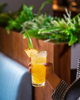 Woman is holding a tropical cocktail in a tiki glass, garnished with a pineapple wedge, in a bar setting