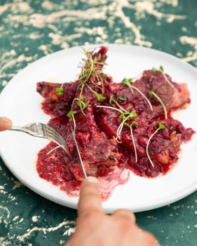 Grilled lamb ribs with cranberry sauce and microgreens are being cut by a chef on a white plate