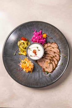 Sliced roast beef with spices is served on a black plate with pickled cucumbers, cabbage and sauce