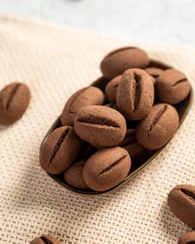 Chocolate cookies shaped like coffee beans are piled in a dish sitting on a beige linen tablecloth