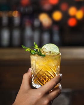 Bartender is holding a refreshing whiskey cocktail with lime and mint against a blurred bar background
