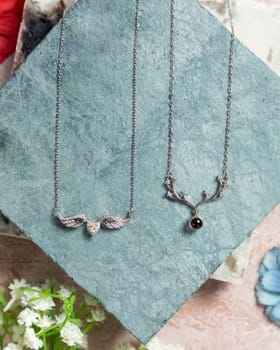 Two silver necklaces on textured stone, surrounded by flowers, showcasing their elegant design