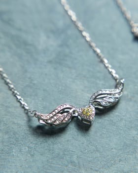 Silver necklace with heart shaped pendant decorated with sparkling diamonds and angel wings is lying on blue background