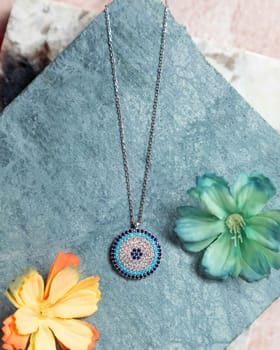 Silver necklace with blue evil eye pendant laying on blue stone background with flowers