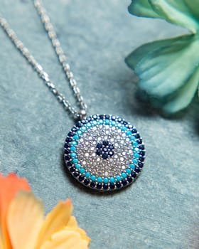 Silver necklace with an evil eye pendant decorated with blue and white crystals lying on a blue background with flowers