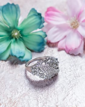 Silver ring with many diamonds lying on a table with two flowers, a green one and a pink one