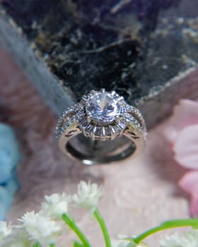 Silver ring with a large center stone and smaller surrounding stones rests on a dark colored stone with small white flowers surrounding