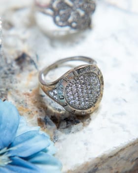 Silver ring with diamonds on stone surface with other rings and blue flower out of focus in the background