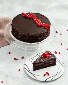 Chocolate cake with mirror glaze decorated with red currant on gray background with a slice of cake on a plate