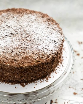 Chocolate cake with chocolate shavings and powdered sugar is sitting on a glass cake stand