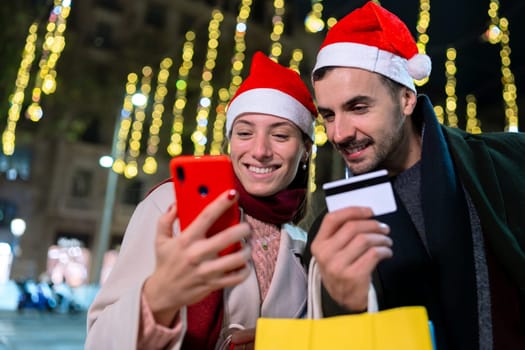 Smiling couple wearing Santa hats, standing under festive Christmas lights. They look at each other warmly, creating a joyful holiday atmosphere. One holds a phone, capturing memories.