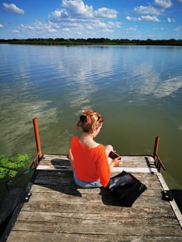 A young woman in an orange top relaxes on a dock by a serene lake, absorbed in her thoughts as gentle ripples reflect the sky and greenery around her.