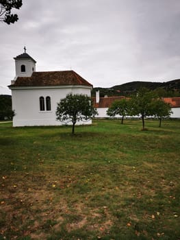 A quaint white chapel stands among green trees, with a serene grassy area around it, under a gray sky in a peaceful rural setting. The atmosphere is calm and reflective.