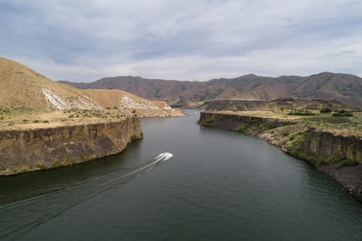 A boat is traveling down a river in a mountainous area. The water is calm and the sky is cloudy