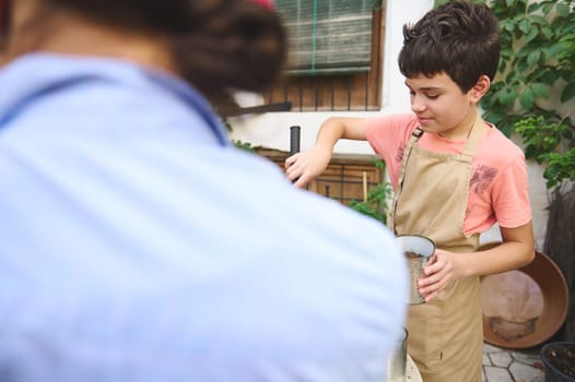 A young boy engaged in gardening outdoors, wearing a beige apron, handling soil in metal pots. The setting is lush and green, evoking a sense of peaceful activity and learning.