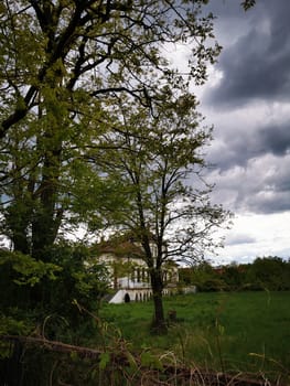 A grand old structure emerges from behind tall trees, surrounded by vibrant foliage and a vast green field, under a moody sky of dark clouds, evoking a tranquil atmosphere.