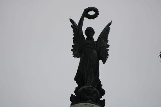 An intricately detailed statue of an angel stands atop a tall column, holding a laurel wreath against a backdrop of a cloudy sky. The scene evokes a sense of reverence and beauty.
