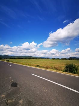 A smooth asphalt road stretches into the horizon, flanked by vibrant green fields and distant mountains. Fluffy white clouds float above in a clear blue sky.