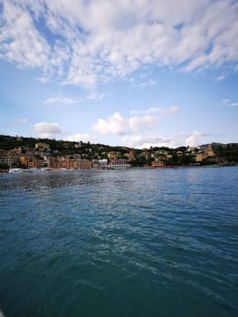 Colorful coastal buildings reflect off the calm water, surrounded by green hills and a bright blue sky, creating a serene seaside atmosphere.