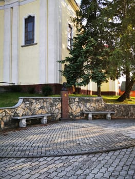 Stone benches provide a tranquil spot in front of a historic building, featuring a decorative bust surrounded by greenery on a bright day.
