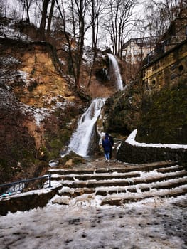 A person walks along a snowy path near a cascading waterfall in a tranquil forested area. The winter scene highlights the natural beauty of the landscape.