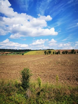The vast landscape shows freshly plowed fields under a bright blue sky filled with fluffy clouds. Green hills rise in the distance, signifying fertile land ready for cultivation.