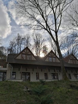 A charming historic house stands amidst tall trees, with sunlight filtering through branches, creating a peaceful atmosphere at dusk.