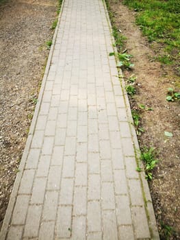 The walkway, made of rectangular stones, runs through a green area, flanked by patches of grass and bare soil, under clear skies.