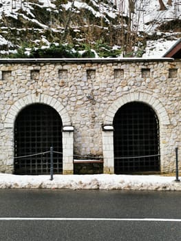 The solid stone structure features two arched entrances surrounded by snow, located beside a quiet roadway in a picturesque mountainous landscape.