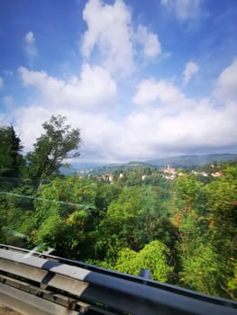 A vibrant landscape unfolds along a highway, showcasing green forests and distant hills beneath a blue sky filled with fluffy clouds.