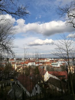 From a high vantage point, the town reveals a blend of rooftops beneath a blue sky filled with fluffy clouds, surrounded by trees and hills in the distance.