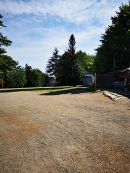 A gravel path leads through a quiet natural area, surrounded by green trees, with hints of an informal gathering space nearby, inviting relaxation and exploration.