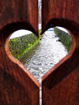 A wooden fence with a heart-shaped cutout provides a charming view of a gentle stream flowing beside lush green moss, showcasing nature's beauty.