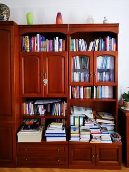 A wooden bookshelf stands in a cozy room, displaying shelves filled with books of various sizes and subjects. Decorative items enhance the inviting atmosphere.