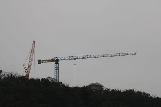 Two construction cranes stand tall against a fog-covered sky, busy overseeing work on a hillside in the early morning. The environment suggests a challenging construction day ahead.