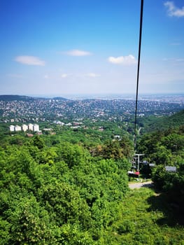 Riding a cable car offers breathtaking views of vibrant green trees and a sprawling city in the distance under a bright blue sky.