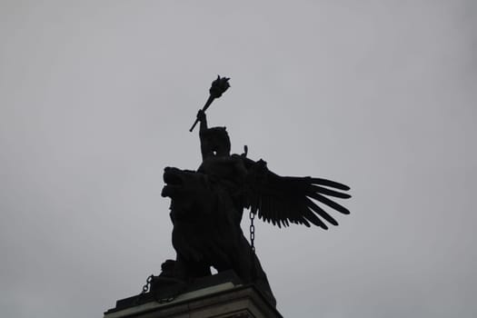 An impressive sculpture depicts an angel with a raised torch, mounted on a lion, silhouetted against a gloomy sky in a serene urban setting.