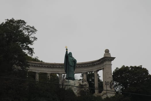 A large statue holding a cross overlooks the landscape, set against a cloudy sky. The structure below enhances the historic ambiance of the site.