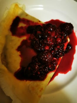 A freshly made crepe is beautifully presented on a white plate, topped with a rich mixture of dark berries and berry sauce, ideal for a sweet breakfast or dessert.