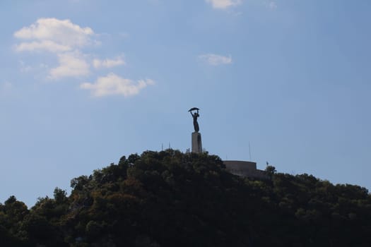 A striking monument of a woman holding an umbrella stands proudly atop a green hill, surrounded by trees, with blue skies and fluffy clouds in the background, indicating a serene daytime atmosphere.