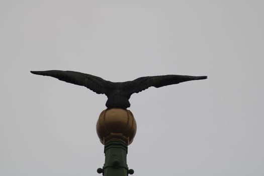 A striking eagle statue stands majestically on a golden sphere, mounted on a green pillar, against a backdrop of overcast skies, showcasing its impressive wingspan.