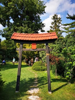 Visitors gather near a traditional wooden torii gate in a lush garden, surrounded by greenery and enjoying a peaceful afternoon under the blue sky.